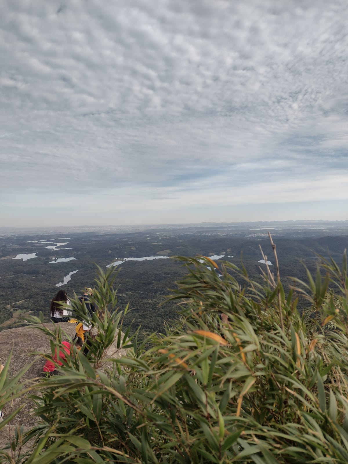 Paisagem de cima da serra, vendo ao fundo uma vista plana e com relevo mais abaixo, um rio e as nuvens no céu e duas pessoas sentadas admirando a vista